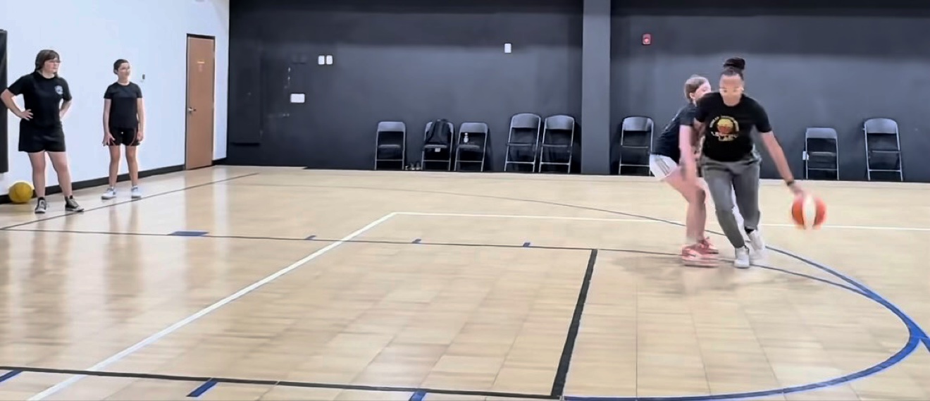 Coach J demonstrating a dribbling move on the court while her players watch and learn